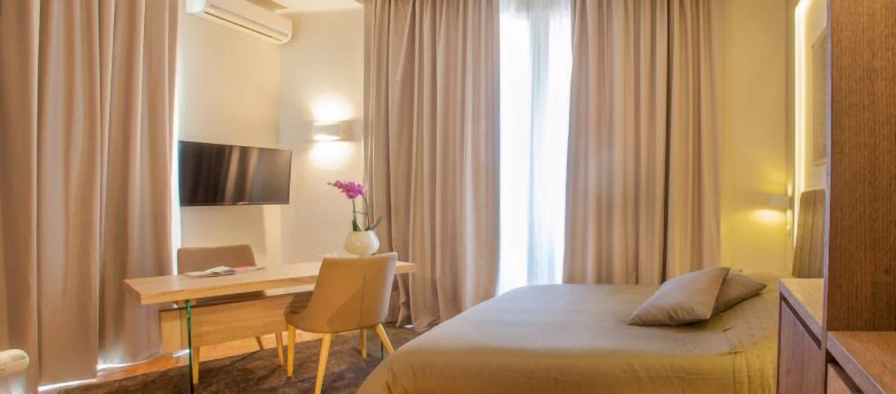 A hotel room with a bed, a desk, a chair, a TV, and a vase with flowers.