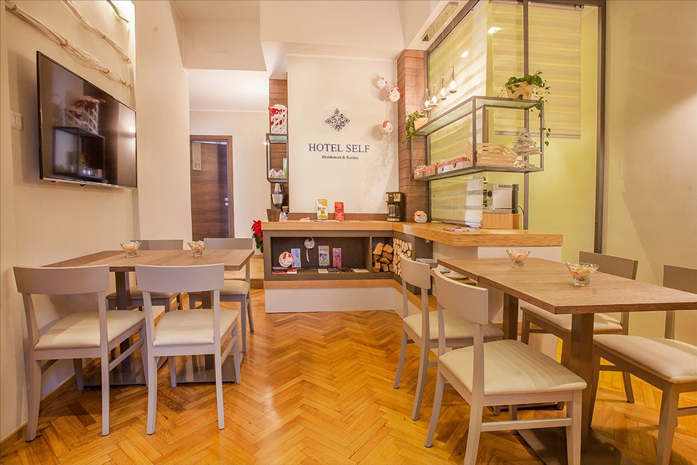 Un espace d’accueil en libre-service dans l’hôtel, avec des tables et des chaises en bois, un comptoir de réception et un téléviseur fixé au mur.