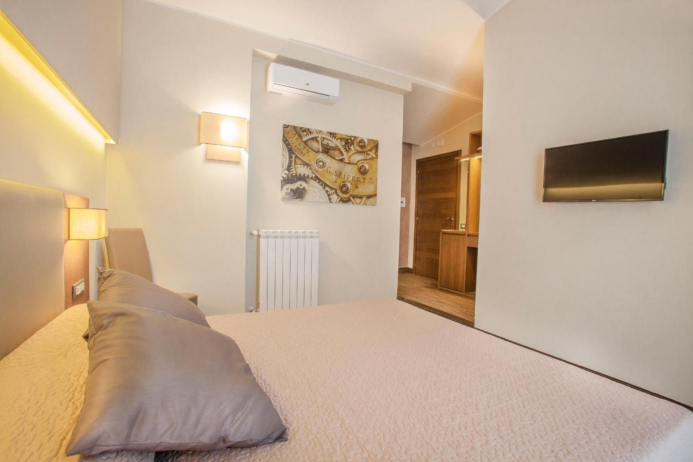 A hotel room with a bed, a wall-mounted TV, a radiator, and a painting.