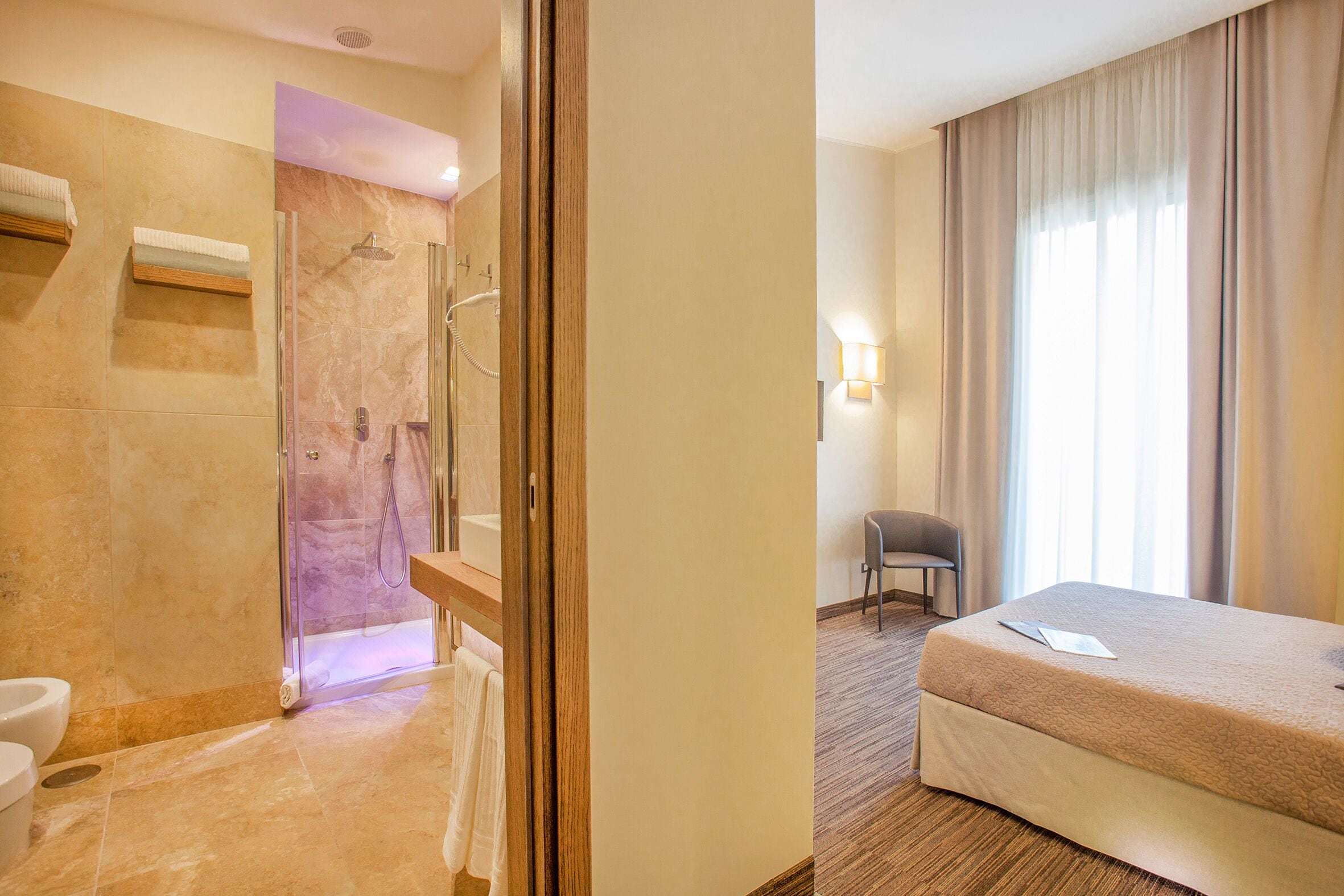 Quarto de hotel com chuveiro, cama, cadeira e janela com cortinas.