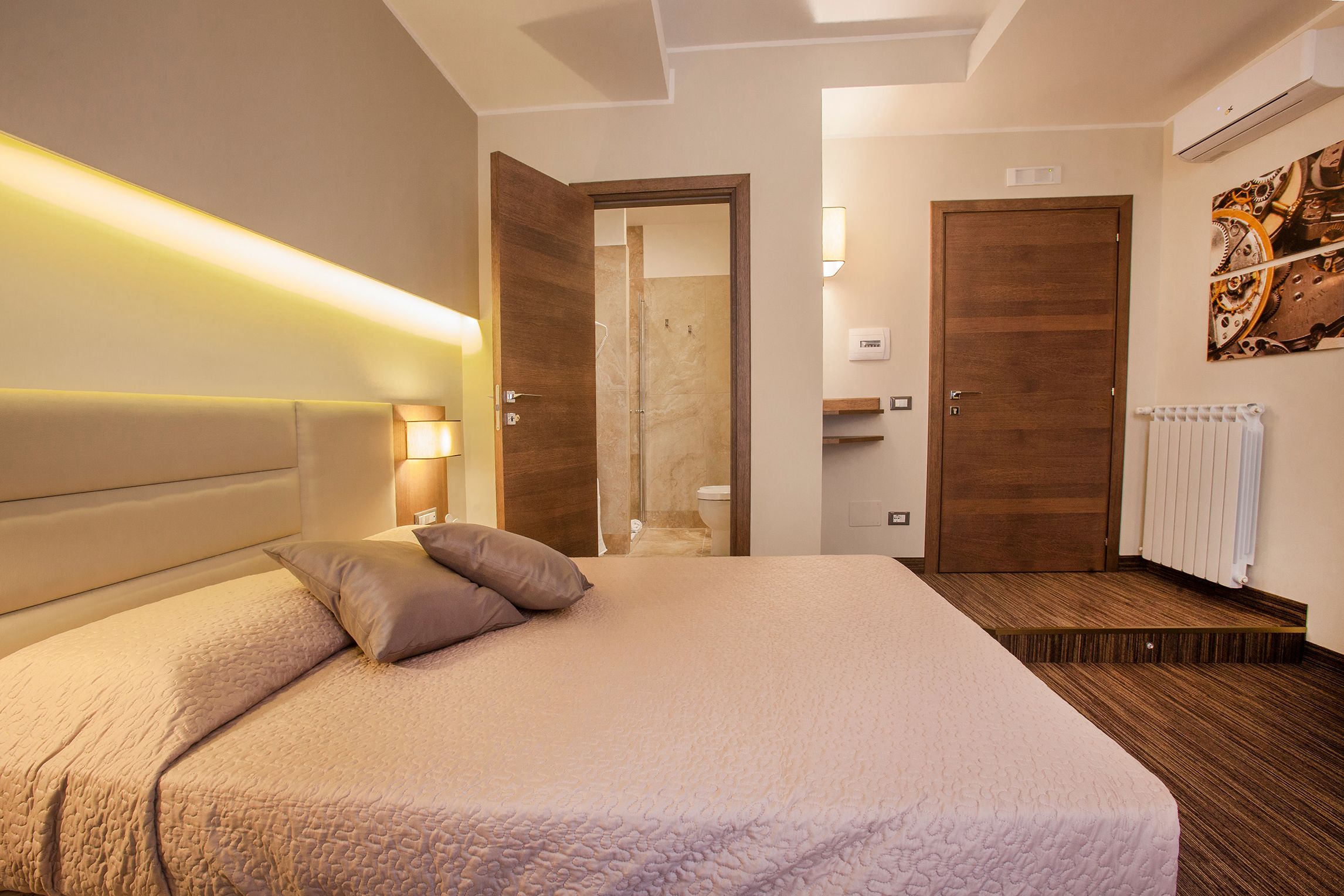 A bedroom with a large bed, a headboard, a nightstand with a lamp, a wooden door, and a bathroom visible through an open door.