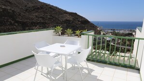 Un balcon avec une table et des chaises blanches, une vue sur la mer et une colline rocheuse.