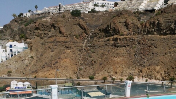 Une vue sur la piscine, avec une assiette de fruits et un sandwich, donnant sur une colline rocheuse parsemée de bâtiments.