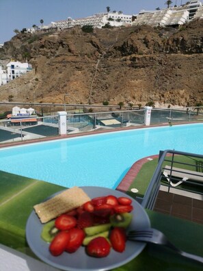 A poolside view with a plate of fruit and a sandwich, overlooking a rocky hillside with buildings. - Apartamentos Halley (Mogan)