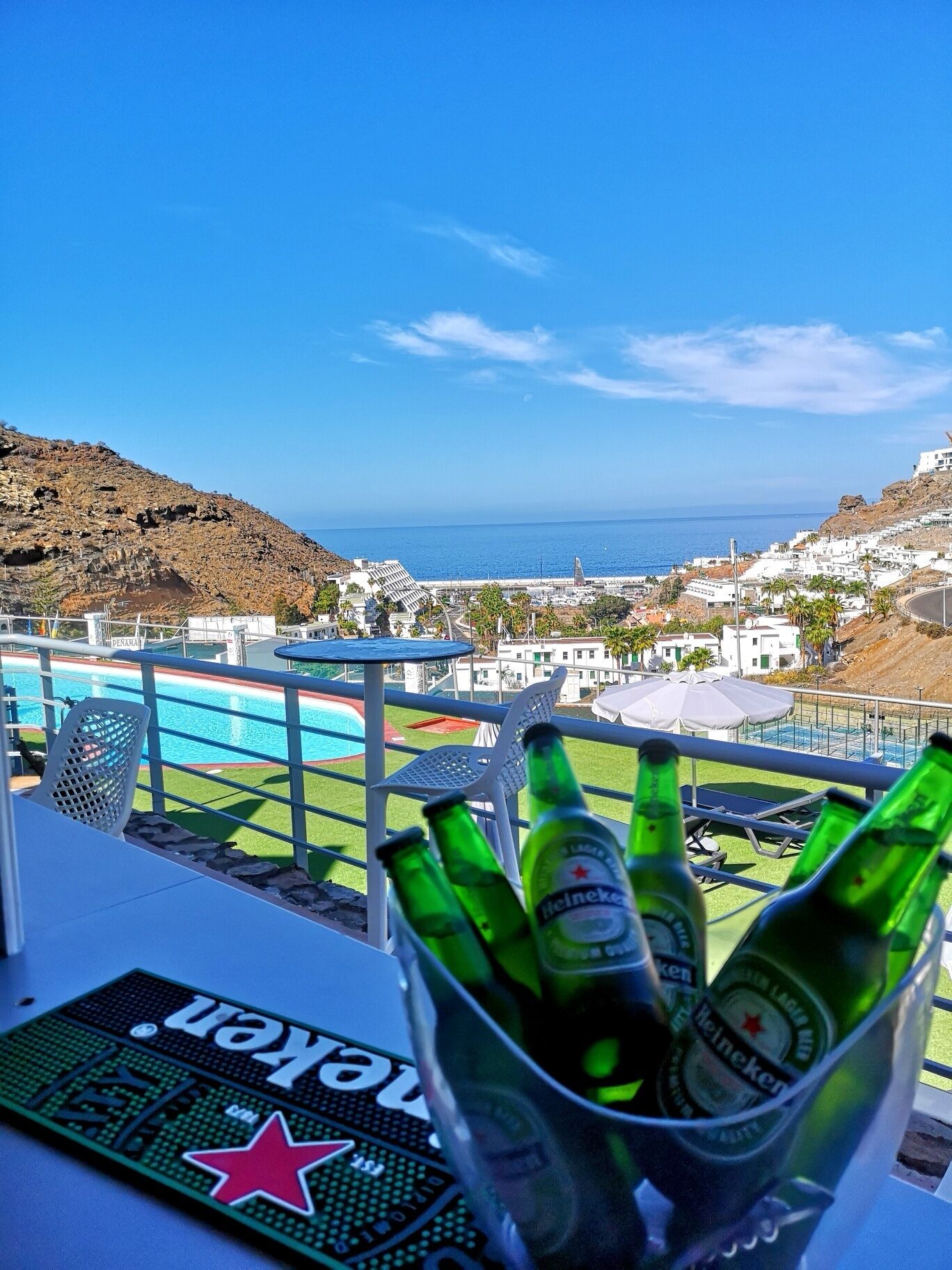 A balcony with a view of the sea, a pool, and several Heineken beer bottles.