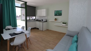 Un salon moderne avec un canapé gris, une table à manger avec des chaises, un coin cuisine doté de meubles blancs et une vue dégagée sur l’extérieur grâce à une grande fenêtre.