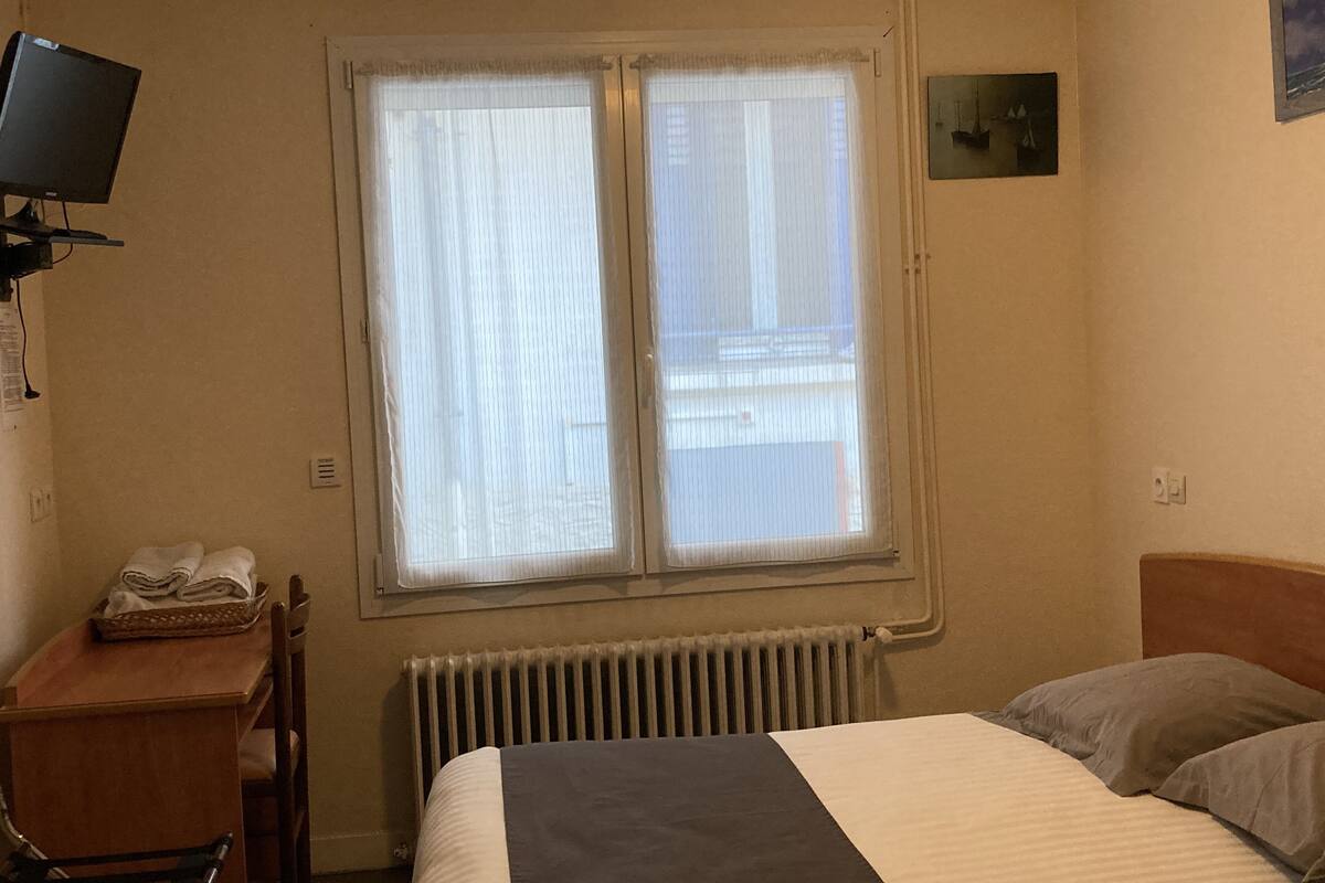 Une chambre d’hôtel avec un lit, un bureau avec un téléphone, un radiateur et une fenêtre avec des rideaux.