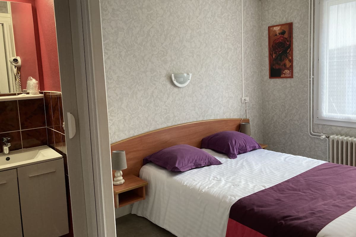 Une chambre à coucher comprenant un lit, une table de chevet, une lampe fixée au mur, un radiateur et une fenêtre avec des stores.