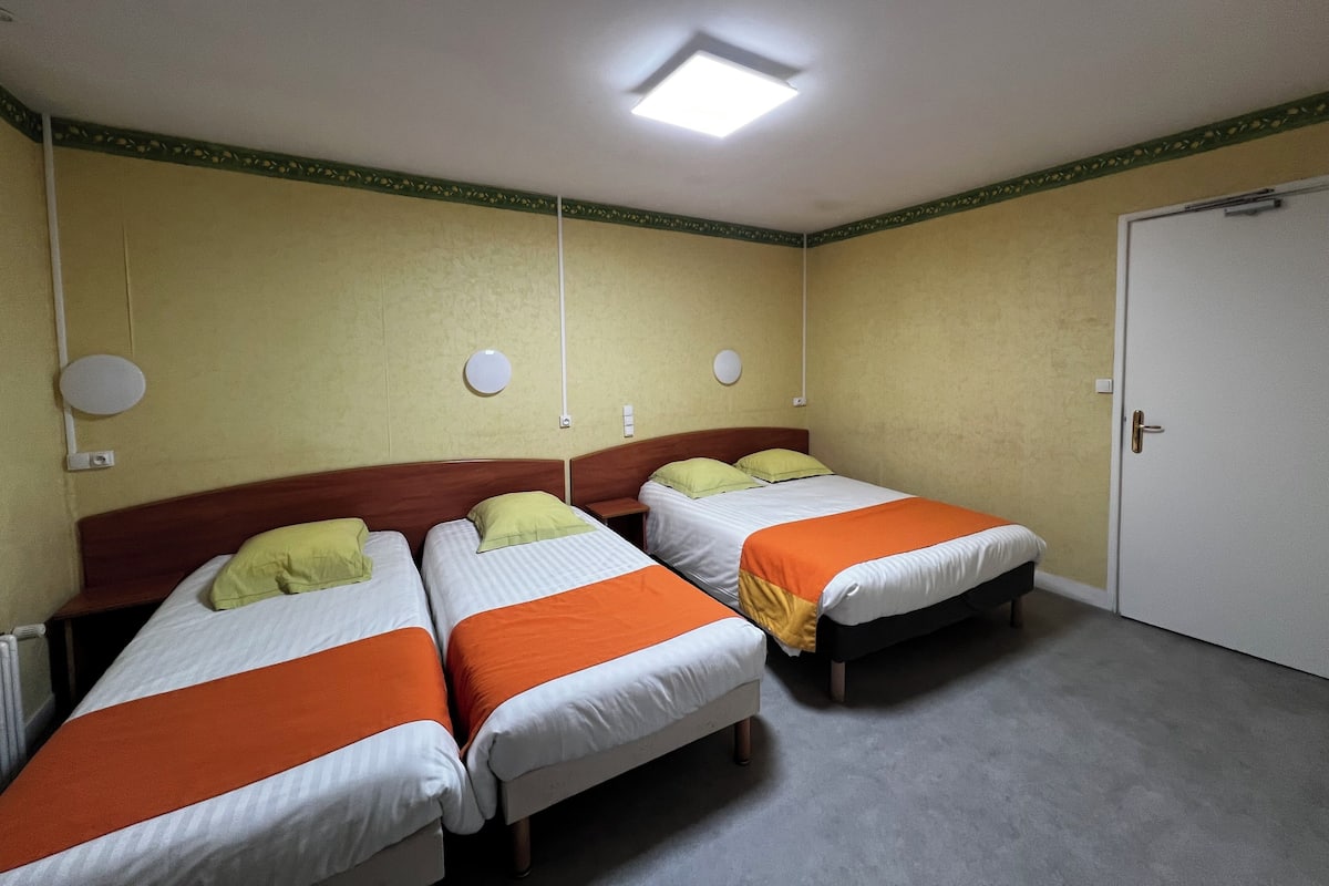 Une pièce avec trois lits, chacun étant équipé d’un oreiller et d’une couverture. Il y a une porte sur le côté droit et un luminaire au plafond.