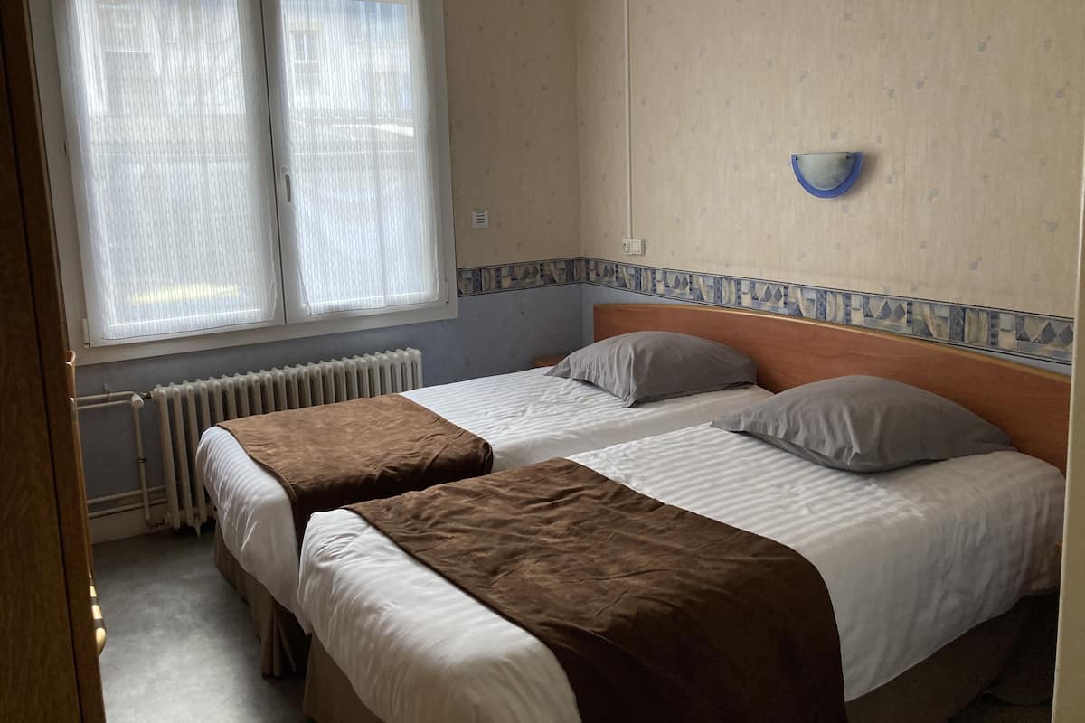 Une chambre avec deux lits, un radiateur, une fenêtre avec des stores et un luminaire fixé au mur.