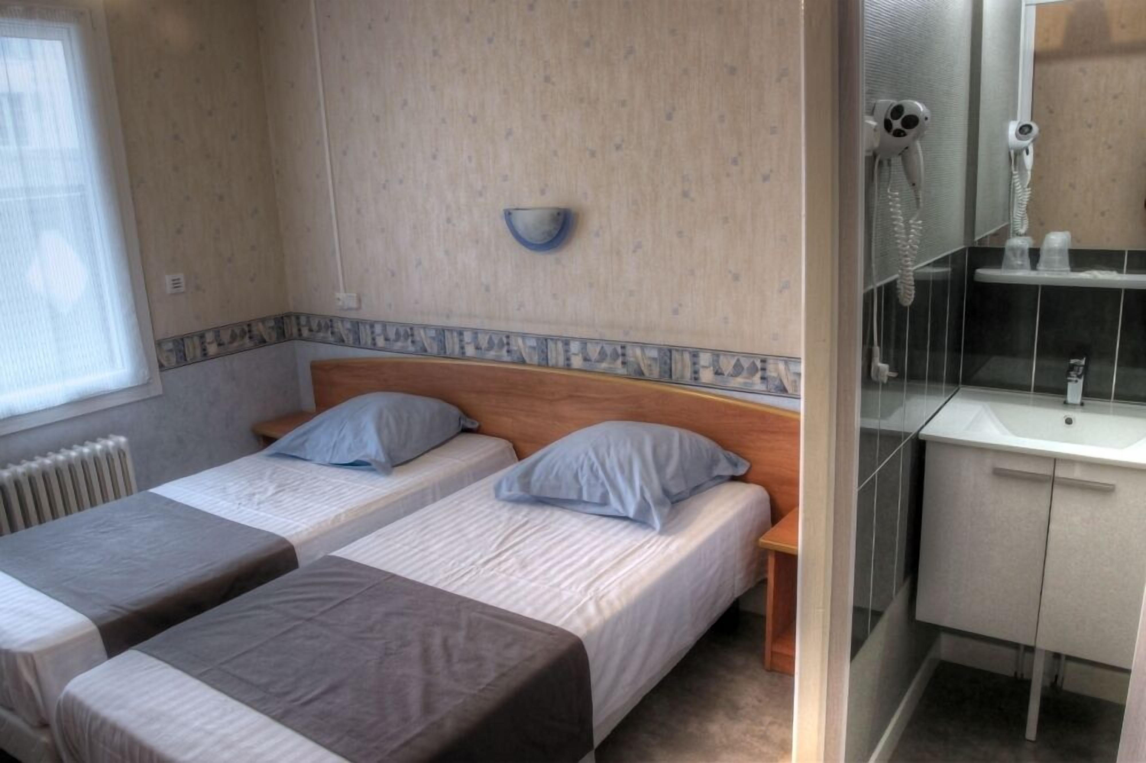 A bedroom with two beds, a small bedside table, a wall-mounted light, and a bathroom with a sink and mirror.