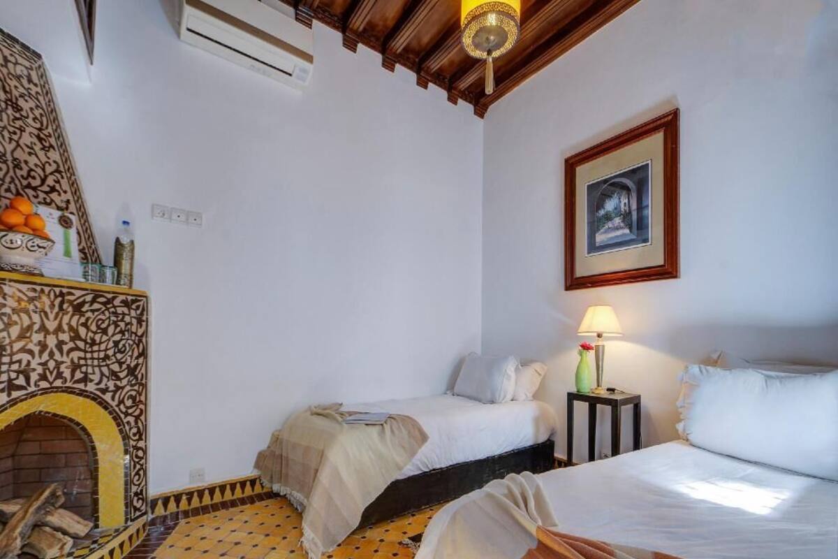 A room with two beds, a fireplace, a framed picture on the wall, and a lamp on a bedside table.