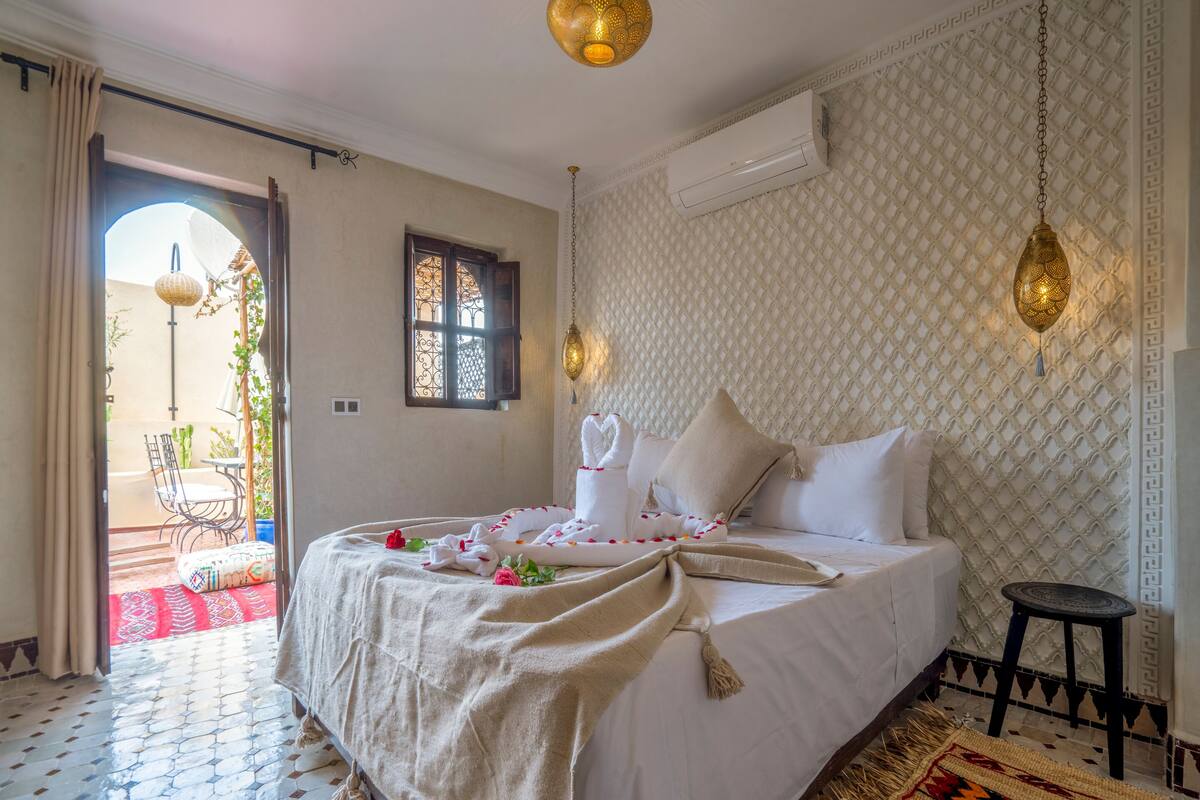 A well-lit bedroom with a bed, a small stool, a decorative wall, and a window.