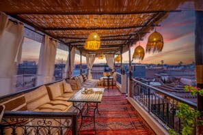 Um terraço acolhedor no telhado, com telhado de palha, assentos estofados, uma mesa pequena com um bule e lanternas penduradas.