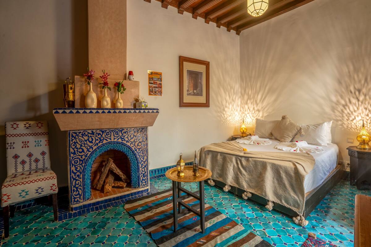 A room with a patterned floor, a fireplace with decorative tiles, a bed with a white bedspread, and a small table with a lamp.