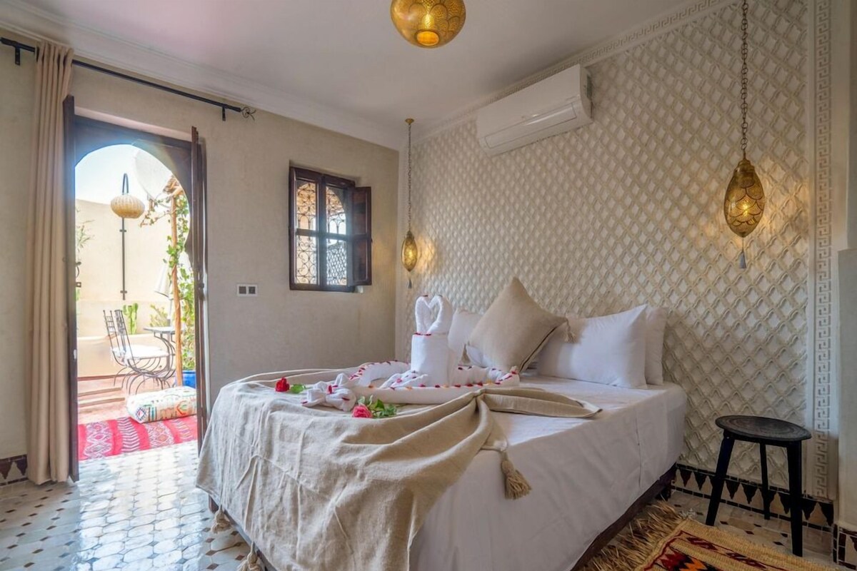A hotel room with a bed, a small stool, a patterned rug, and a view of the outside.