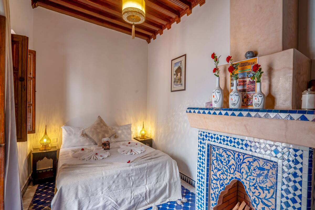 A bedroom with a bed, a nightstand, a wall-mounted lamp, a decorative shelf with vases, and a tiled fireplace.
