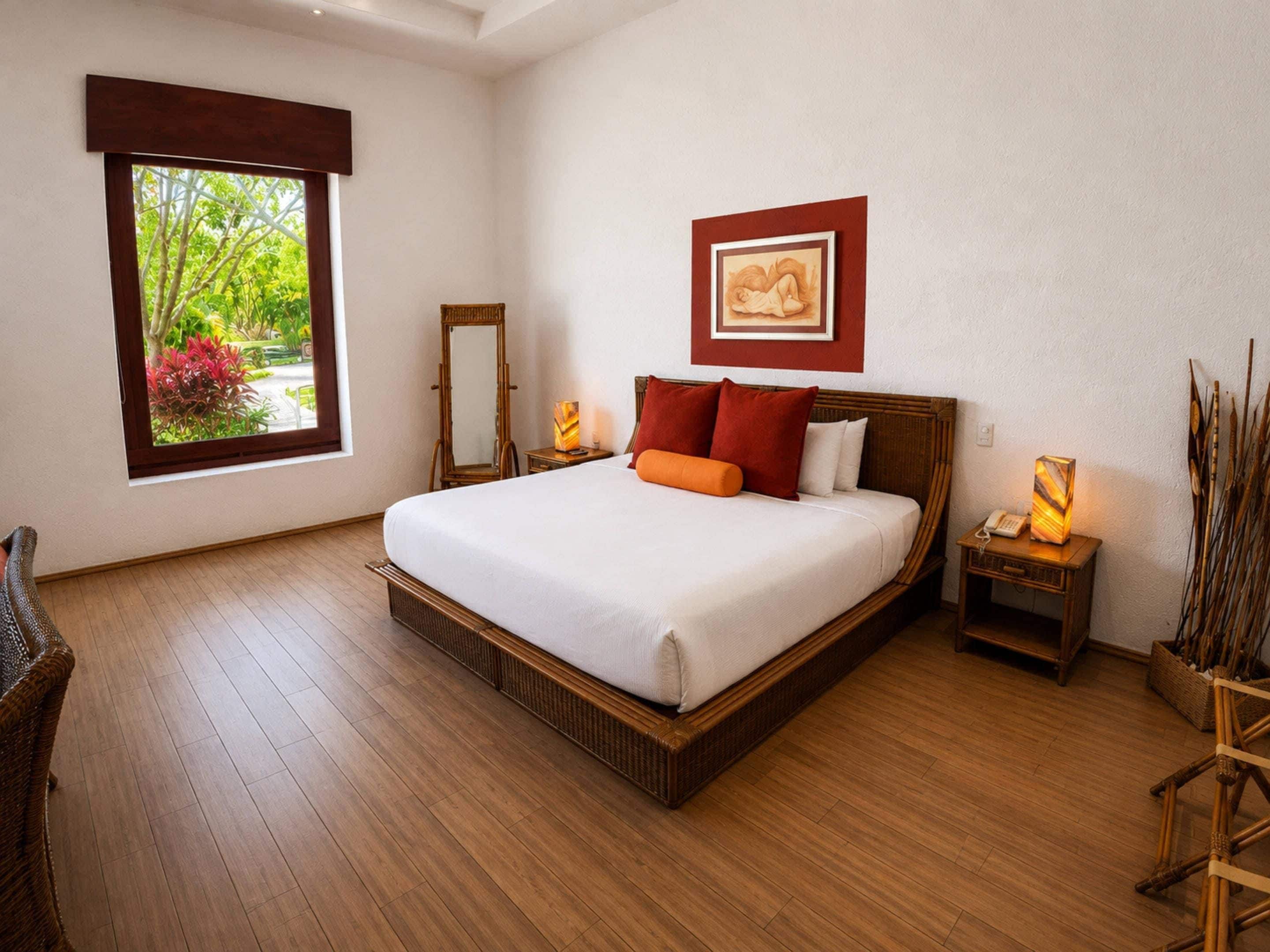 A bedroom with a large bed, a bedside table with a lamp, a mirror, and a window with a view of greenery.