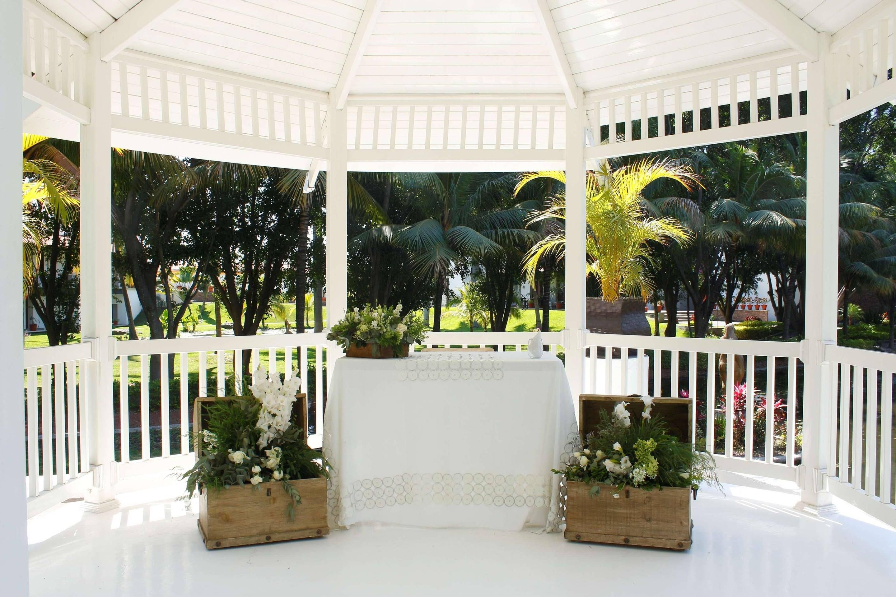 A white gazebo with a white table and benches, surrounded by lush greenery.