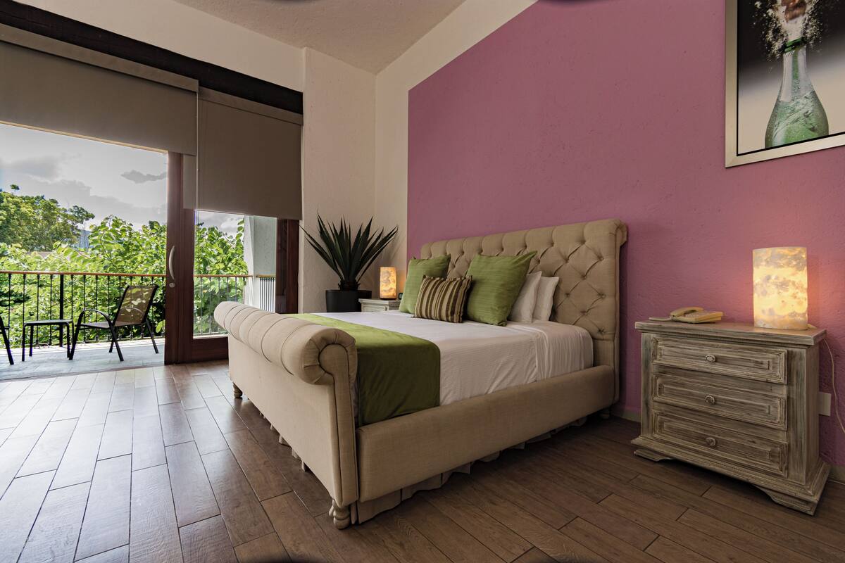 Une chambre avec un grand lit, une table de chevet, une plante en pot et une vue sur l’extérieur.