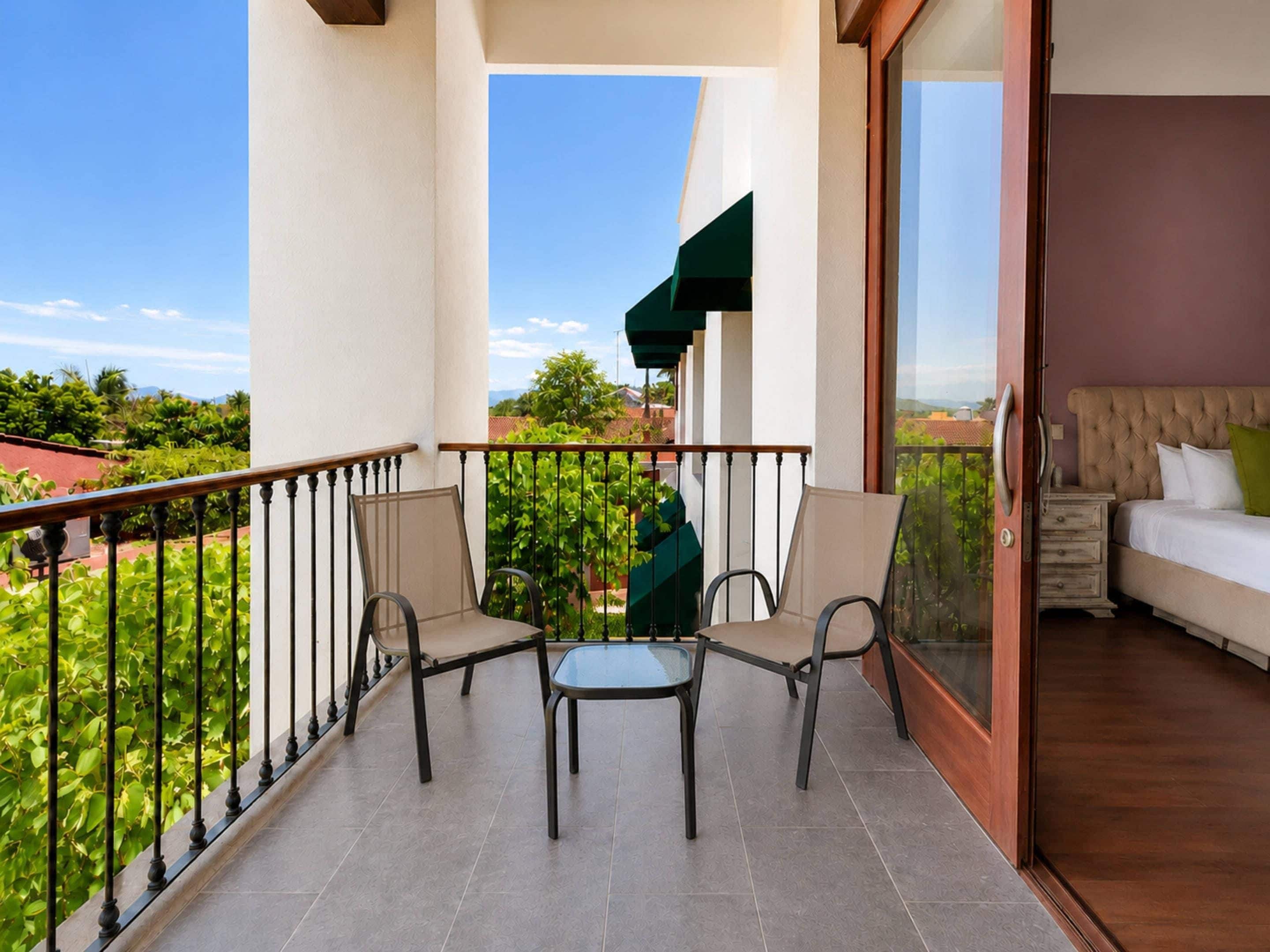 A balcony with a table and chairs, a bed with a green pillow, and a view of greenery and buildings.