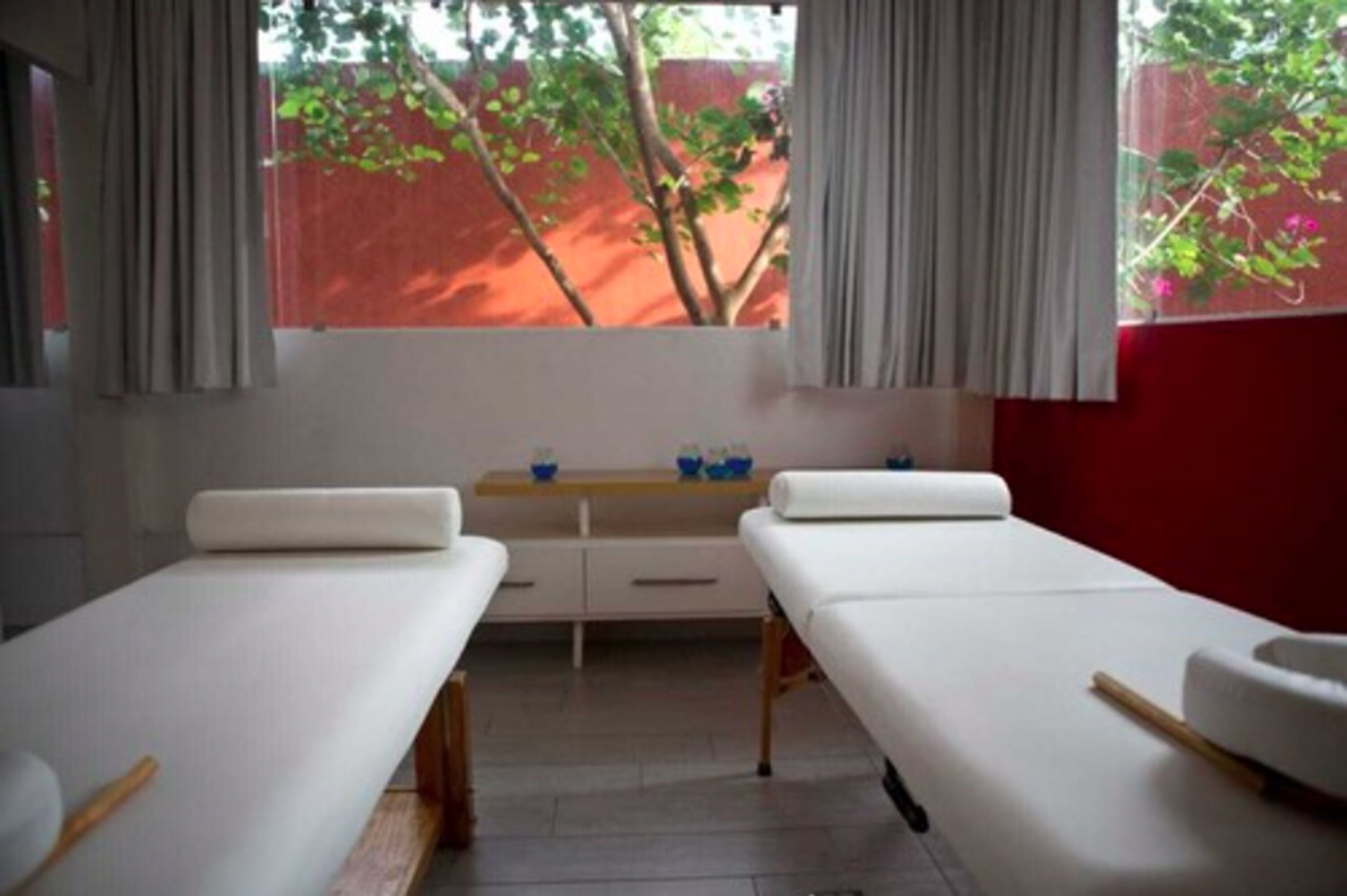 Una habitación con dos camas, un banco de madera, una mesita con cuencos azules y una ventana con cortinas.