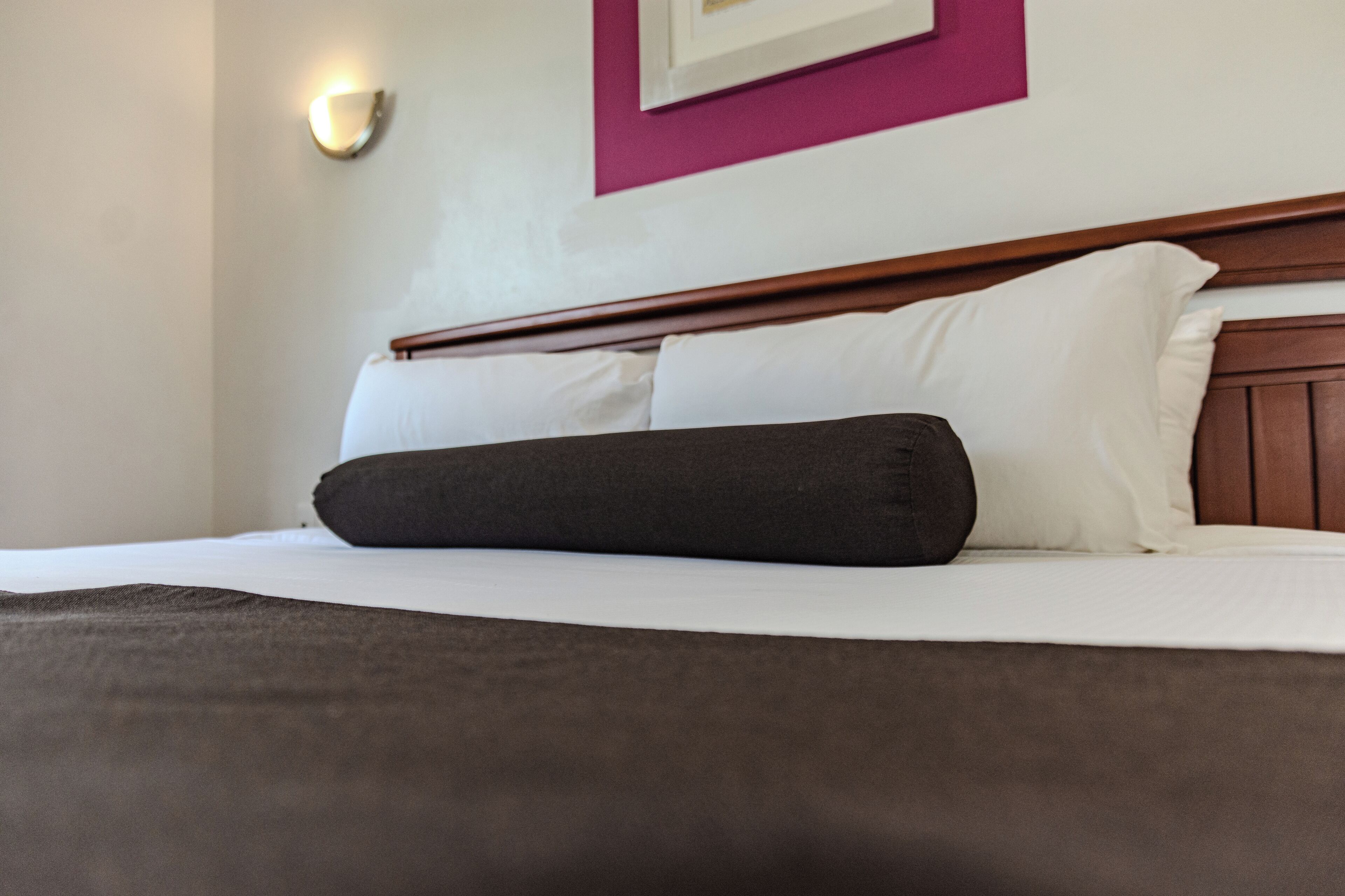 Una cama bien tendida con ropa de cama blanca y marrón oscuro, un cabecero de madera y una lámpara de pared.