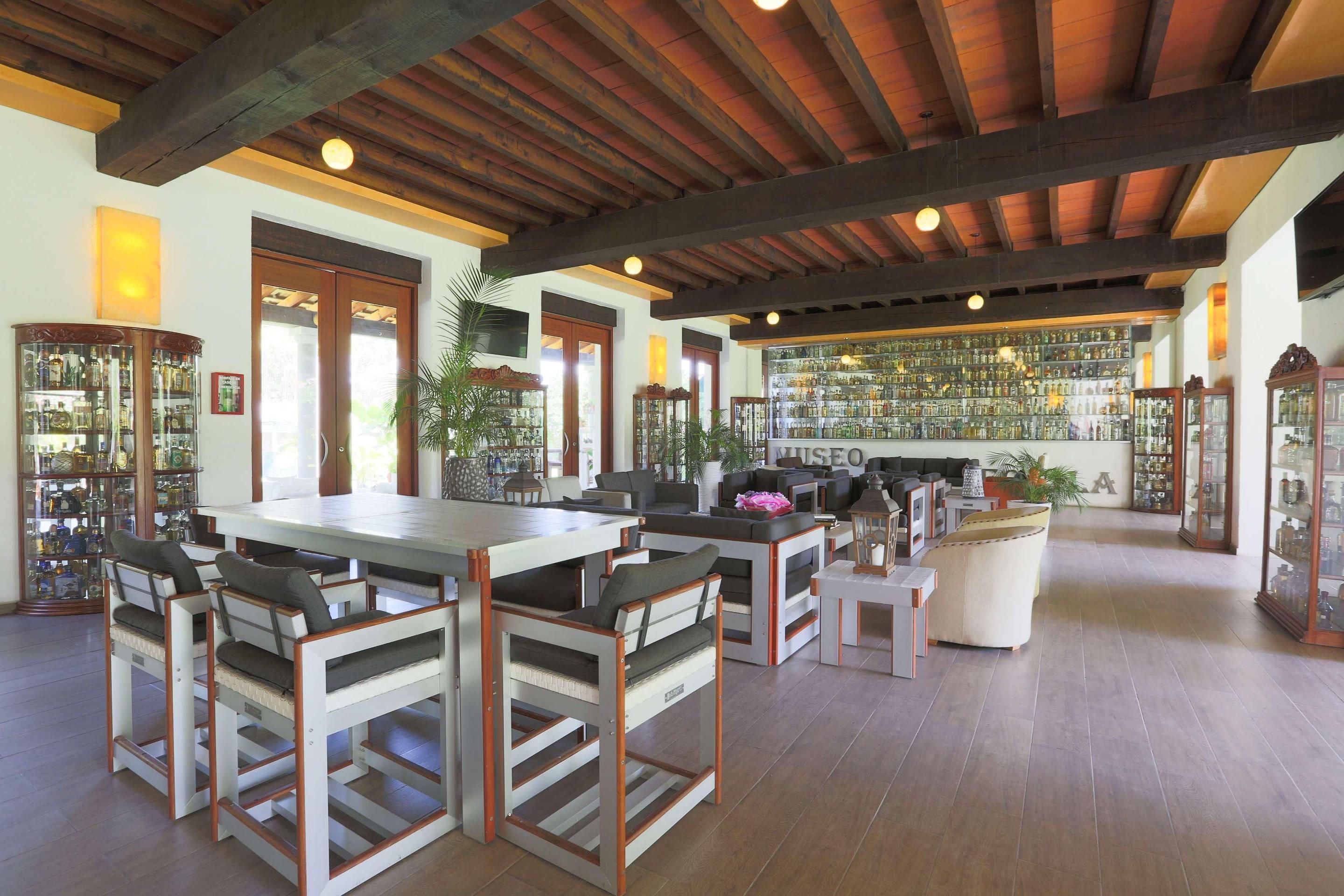 A spacious room with a high ceiling, wooden beams, a central dining table with chairs, and a bar area with shelves of bottles.