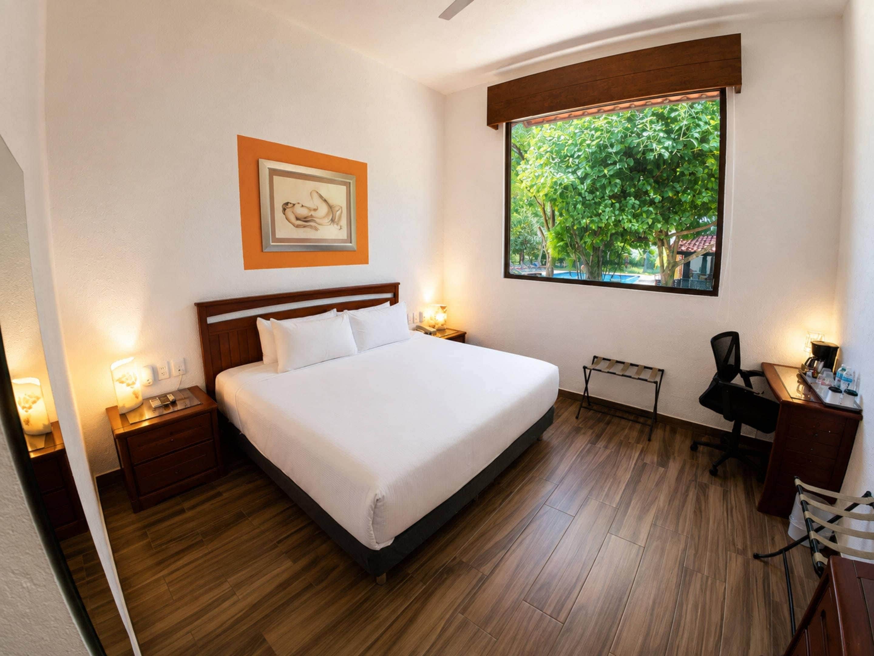 Une chambre d’hôtel avec un grand lit, des lampes de chevet, un bureau et une fenêtre donnant sur des arbres.