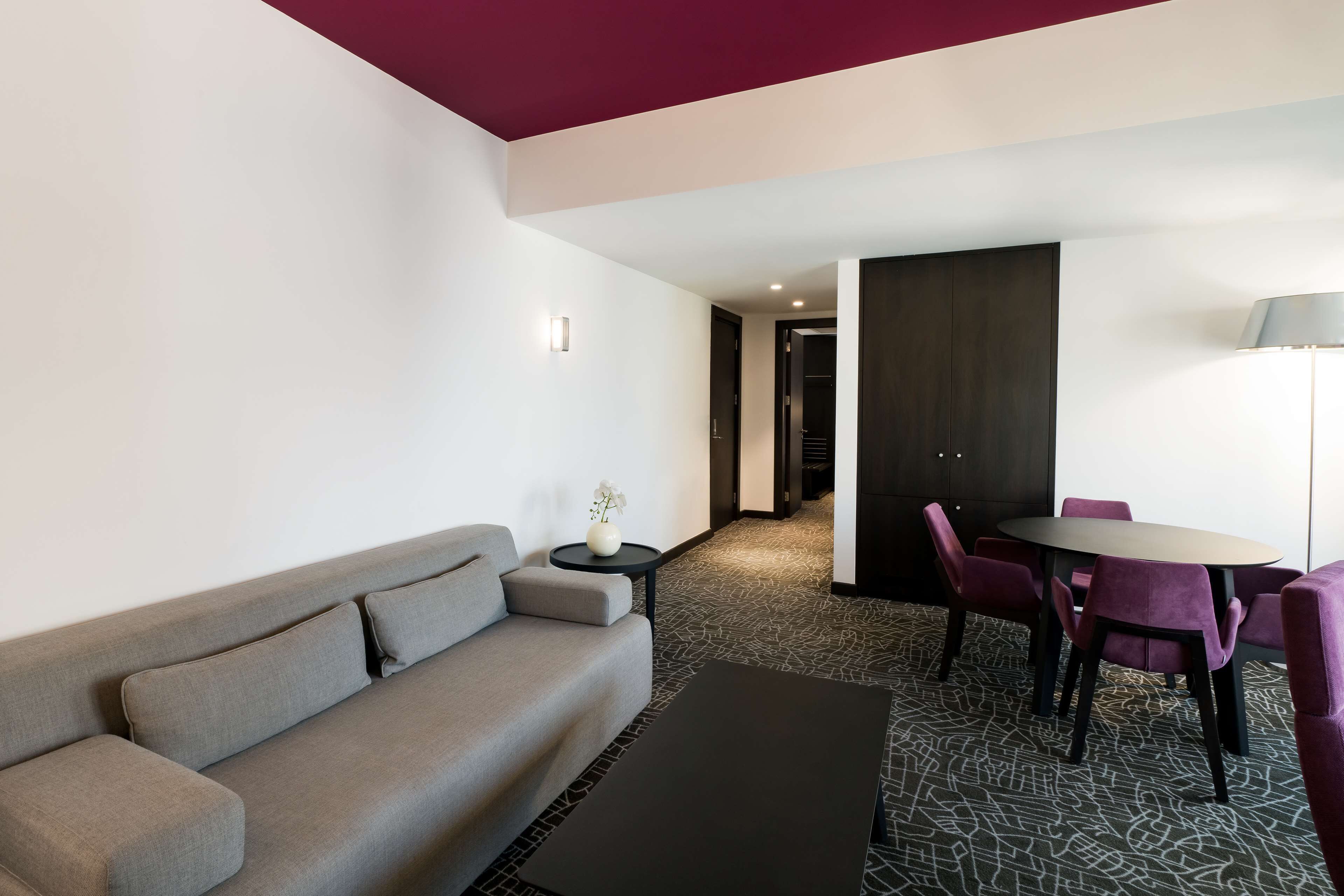 Een moderne hotelkamer met een grijze bank, een kleine ronde tafel met een vaas, een eettafel met paarse stoelen en een kamerbreed tapijt met dessin.