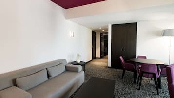 Een moderne hotelkamer met een grijze bank, een kleine ronde tafel met een vaas, een eettafel met paarse stoelen en een kamerbreed tapijt met dessin.