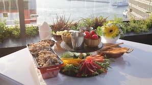 Une table de buffet garnie de divers plats et agrémentée d’un arrangement floral, le tout placé devant une vue sur la mer et un bâtiment.