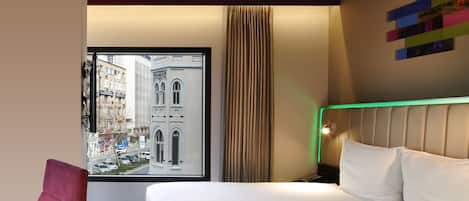A hotel room with a bed, a red chair, a window with a city view, and a wall with a colorful abstract art piece.