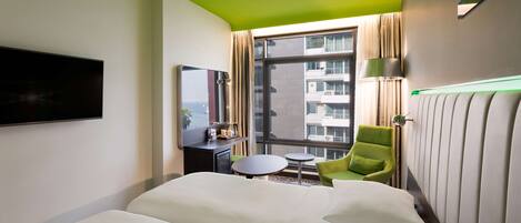 Habitación de hotel con una cama grande, una mesita redonda pequeña, un sillón verde, un televisor montado en la pared y una ventana con cortinas.
