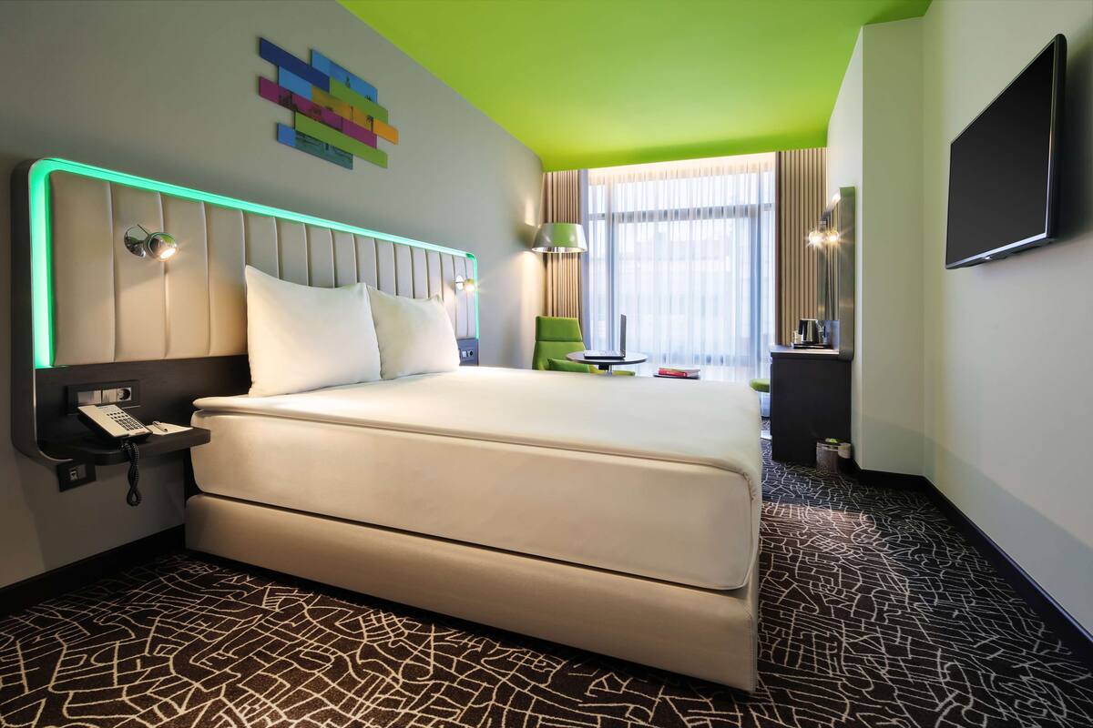A modern hotel room with a large bed, a desk with a phone, a TV, and a green accent wall.