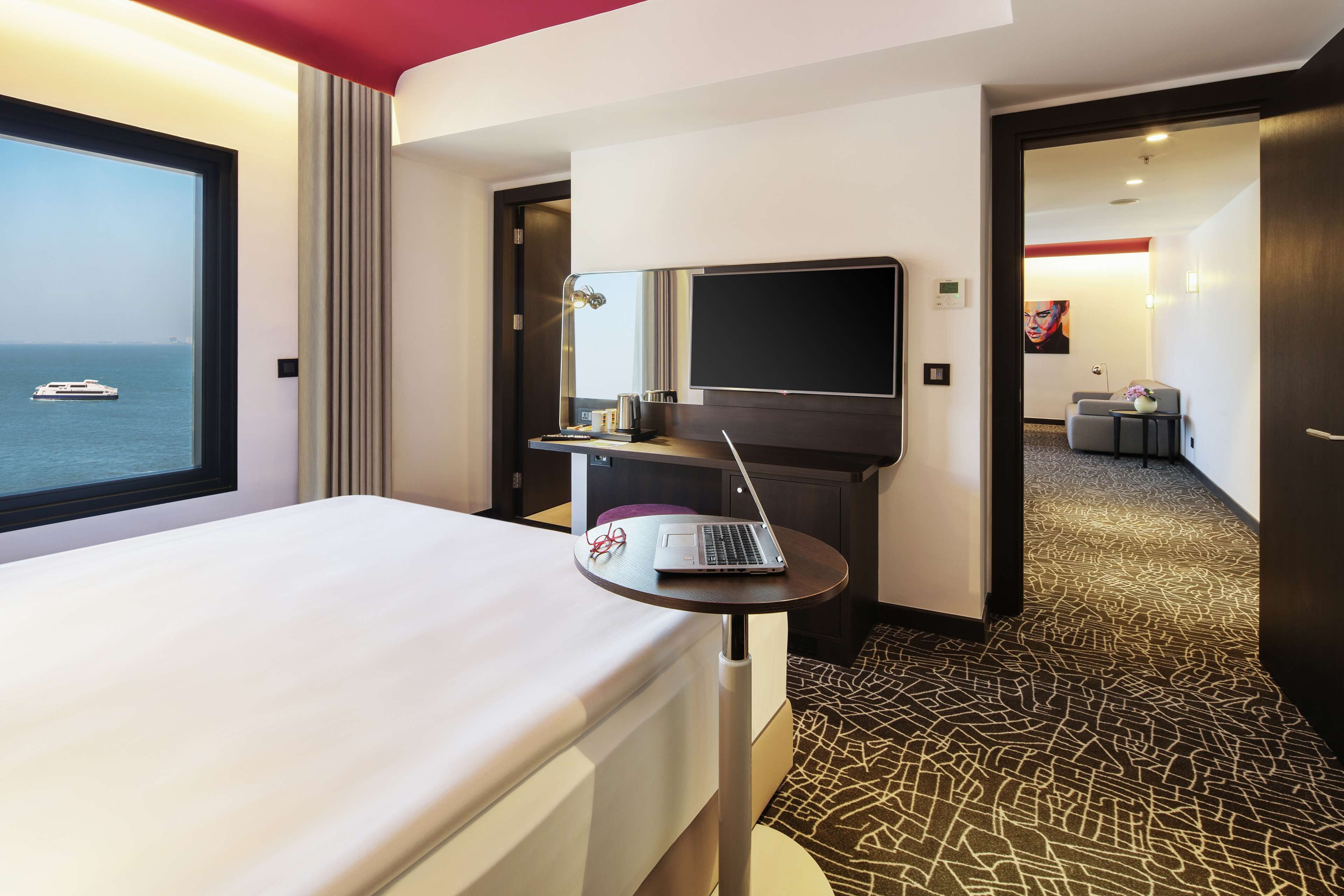 A hotel room with a bed, a laptop on a small table, a TV, and a view of the sea.