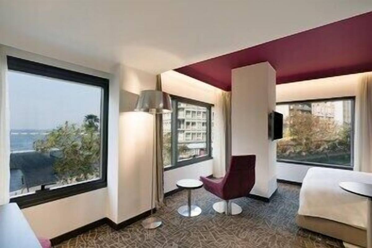 A hotel room with large windows, a flat-screen TV, a red chair, a small round table, and a sofa.