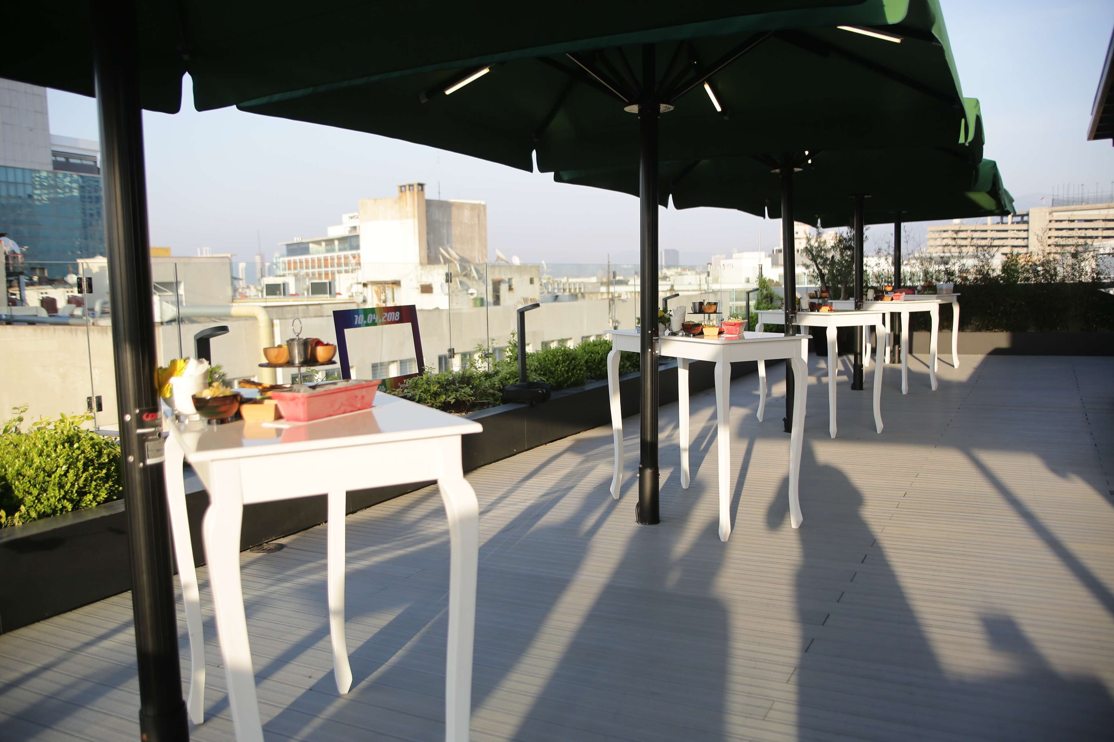 A rooftop terrace with white tables and chairs, a green umbrella, and a view of the cityscape.