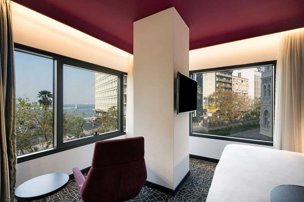 A hotel room with a large window, a flat-screen TV, a red chair, a white bed, and a small round table.