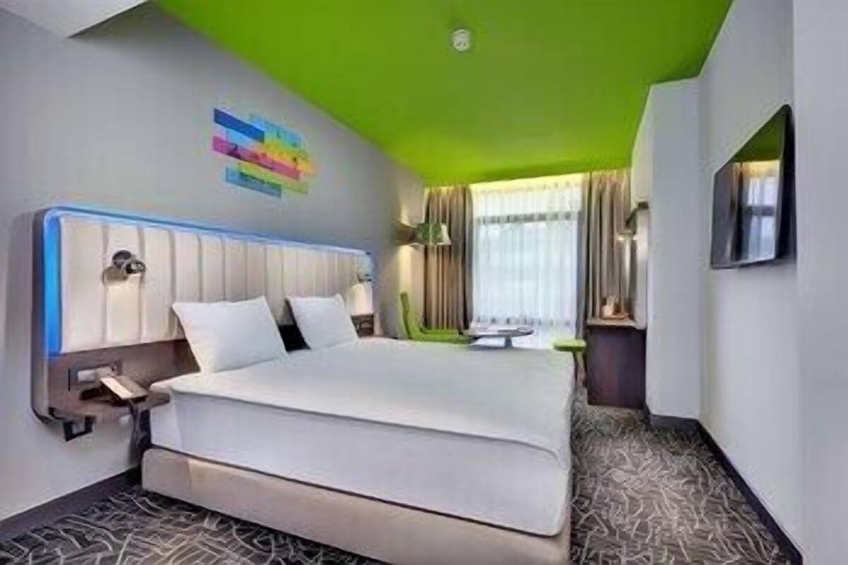 A hotel room with a bed, a nightstand, a lamp, a window with curtains, and a green ceiling.