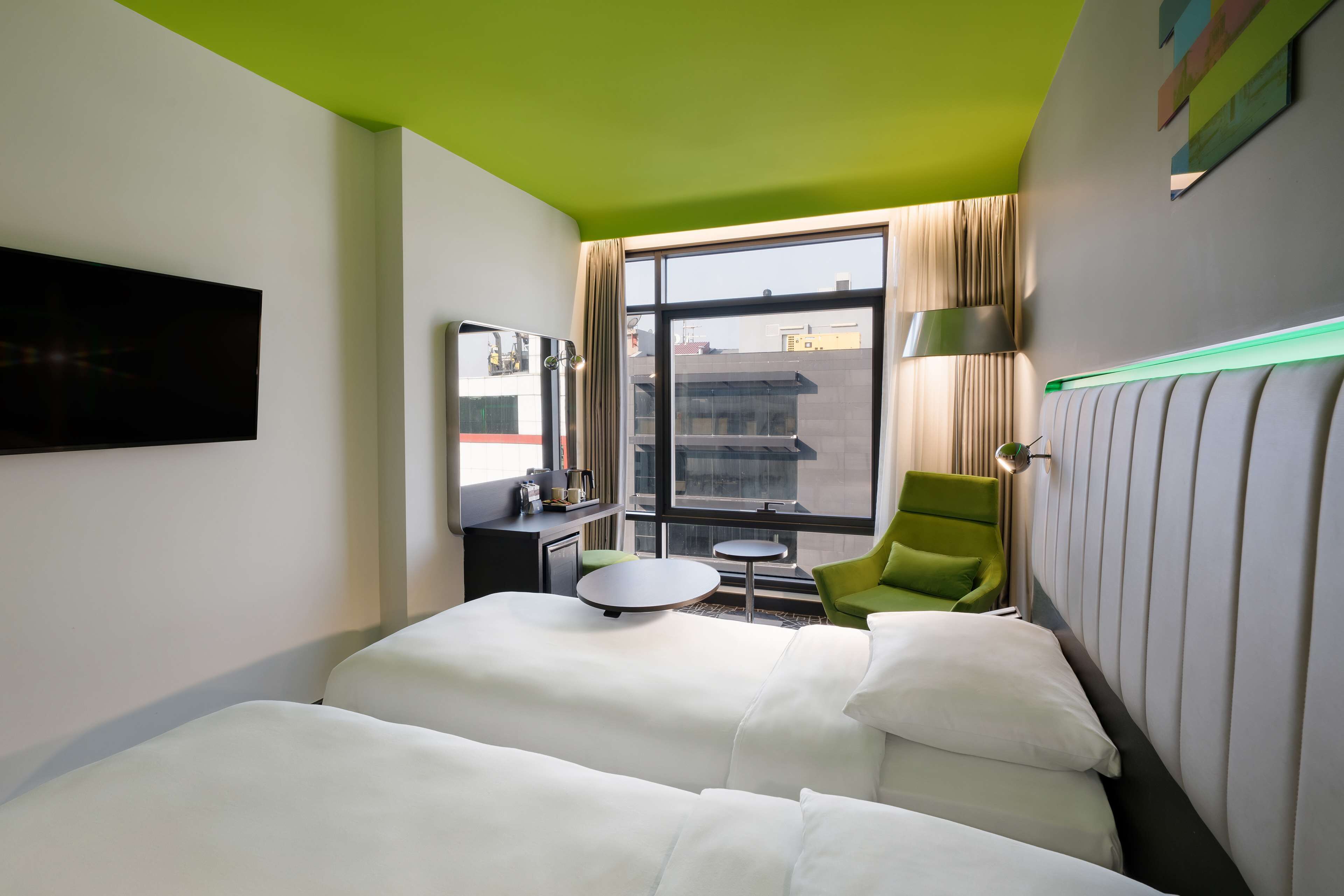 A hotel room with a bed, a small table, a chair, a TV, and a window with a view of buildings.