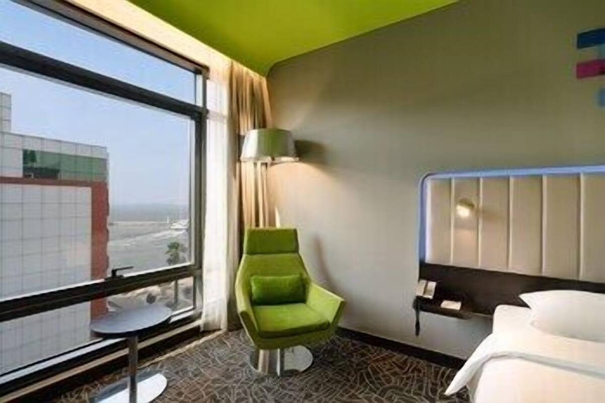 A hotel room with a large window, a green chair, a white bed, and a small table.