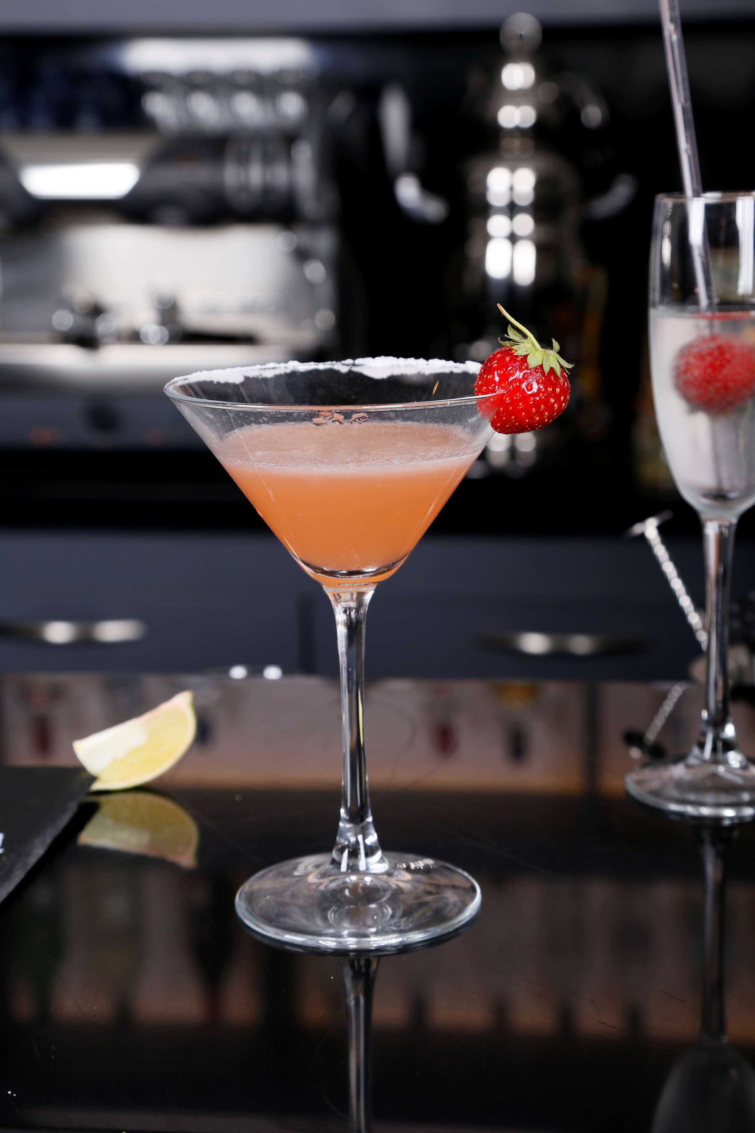 A cocktail in a martini glass with a strawberry garnish, placed on a reflective surface.