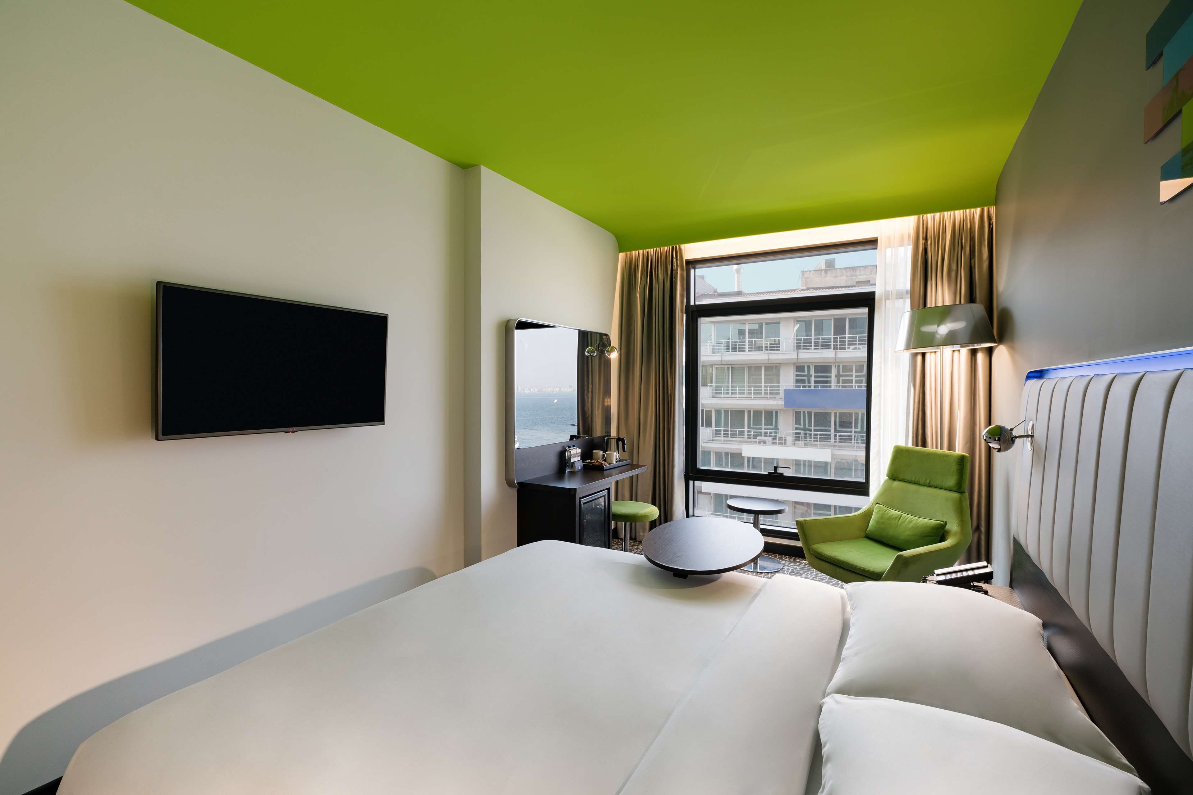 A hotel room with a large bed, a flat-screen TV, a desk with a lamp, a green chair, and a view of a building through the window.
