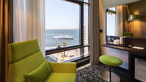 Une chambre d’hôtel dotée d’une grande fenêtre donnant sur la mer et sur un navire de croisière. On y trouve une chaise verte et un petit tabouret près de la fenêtre.