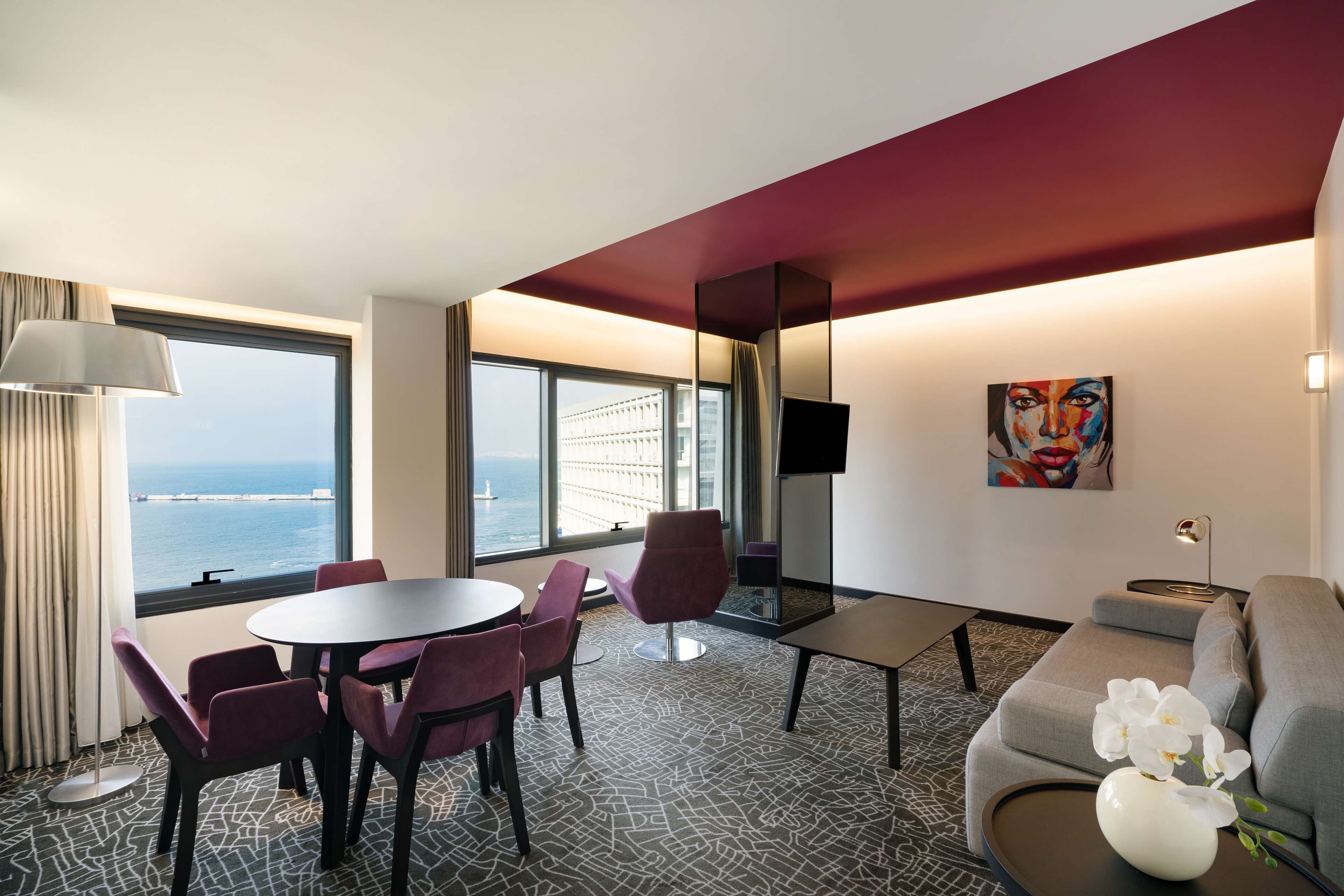 Habitación de hotel con comedor, sofá, ventana grande con vista al mar y un cuadro en la pared.