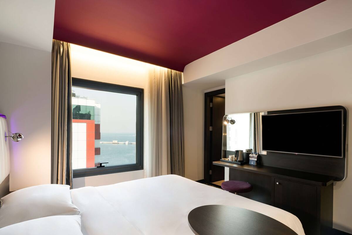 A hotel room with a bed, a desk with a flat-screen TV, a window with curtains, and a view of the city.
