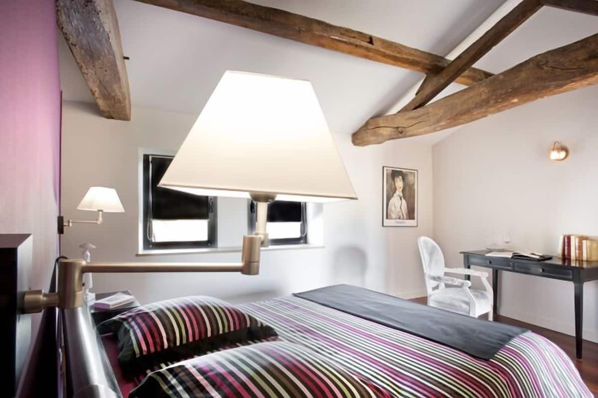 A bedroom with a bed, a desk, a chair, a lamp, and a window.