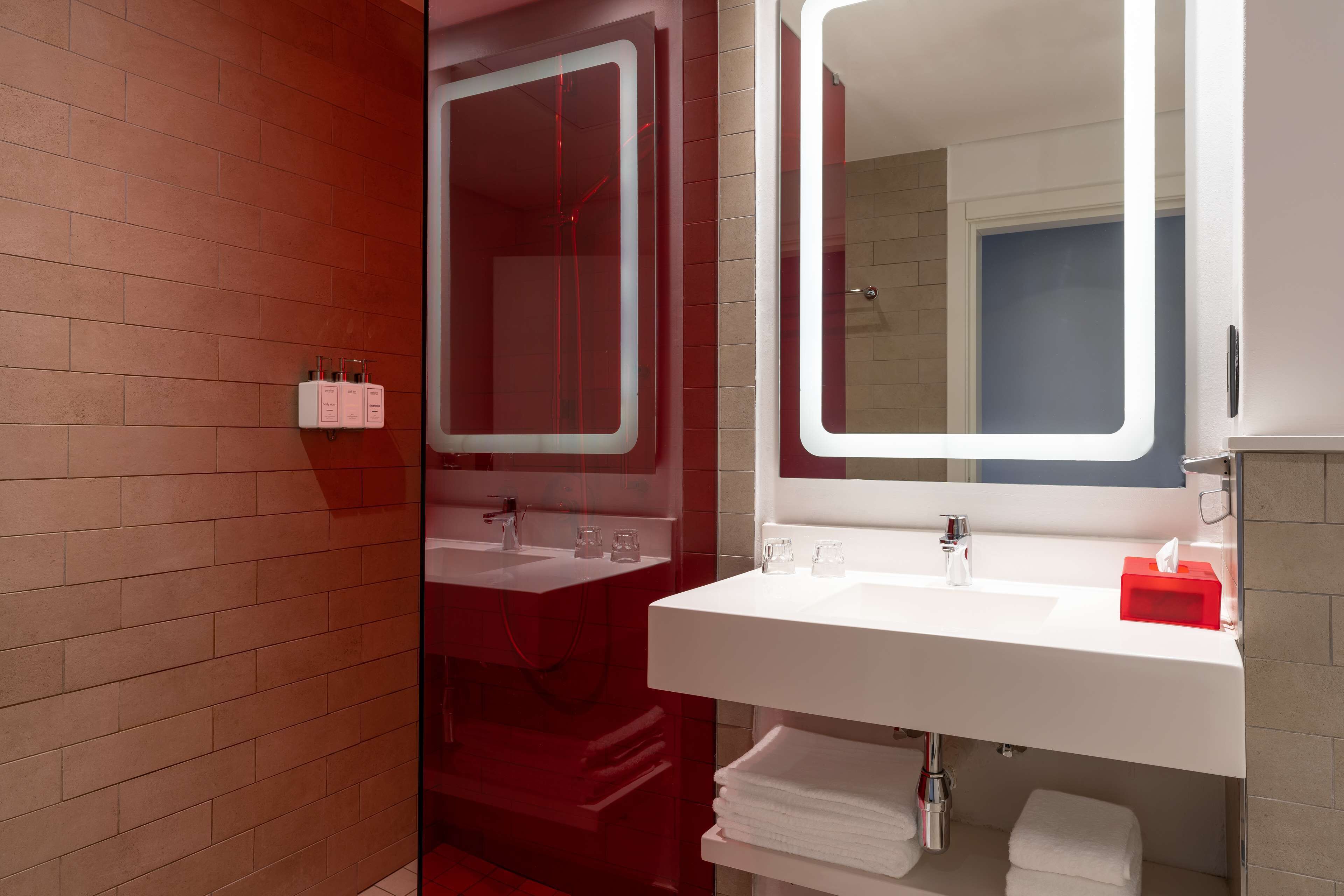 A modern bathroom with a red brick wall, a white sink, a large mirror, and a red soap dispenser.