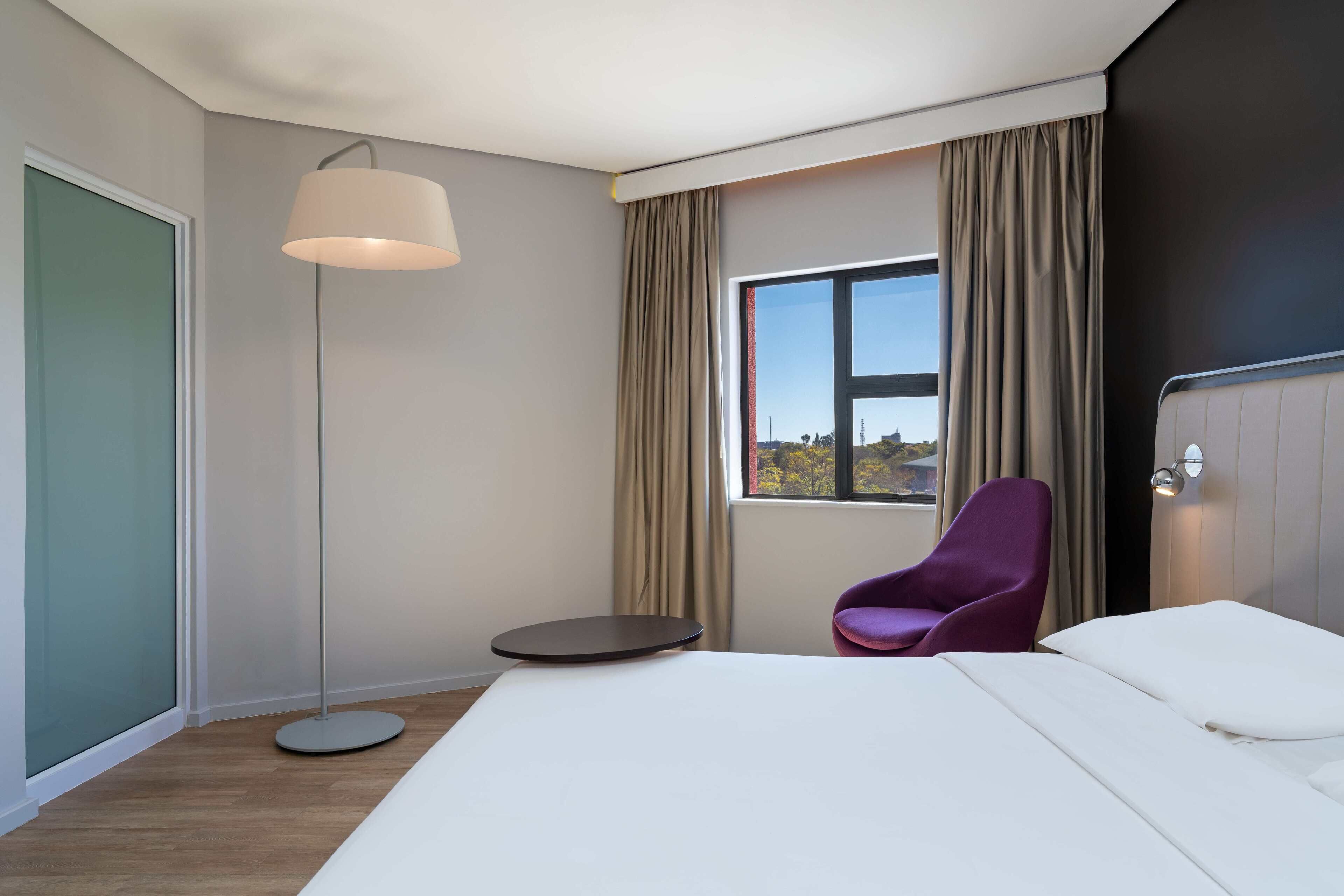 A modern hotel room with a large bed, a floor lamp, a small table, a purple chair, and a window with a view.